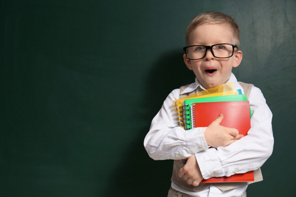 Young boy wearing glasses, holding books and standing in front of a chalk board.